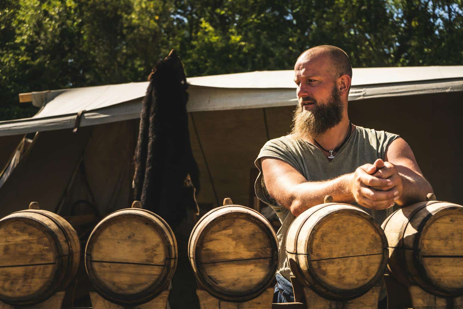 Un homme robuste avec une barbe se tient près de tonneaux en bois dans un cadre extérieur rustique.