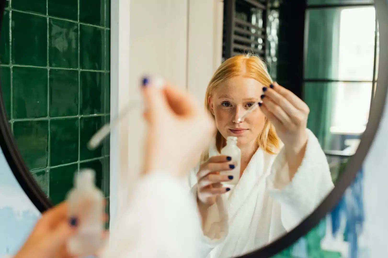 Femme adulte appliquant du sérum de soin tout en portant un peignoir devant un miroir de salle de bain.